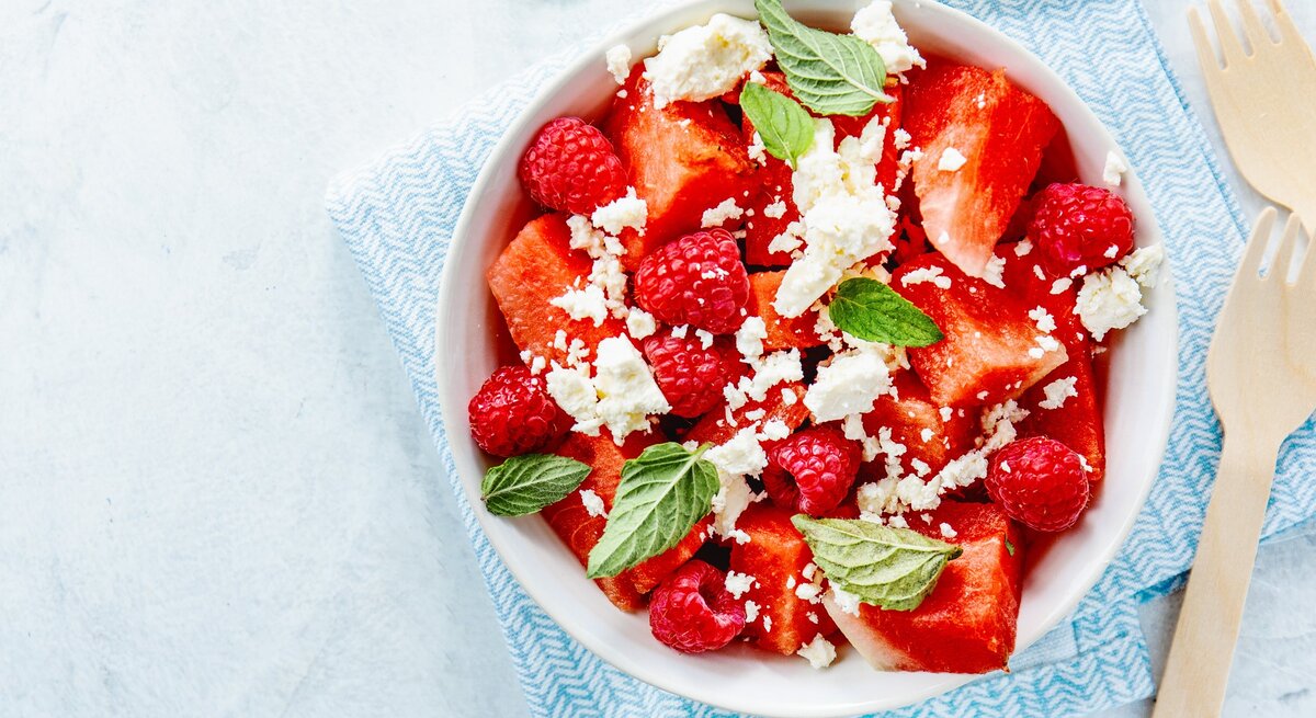 Wassermelonenslata mit Minze, Himbeeren und Feta in einer weißen Schale auf einem blauen Tuch. Auf einem hellen Tisch mit zwei Holzgabeln an der rechten Seite.