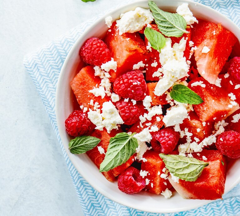 Wassermelonenslata mit Minze, Himbeeren und Feta in einer weißen Schale auf einem blauen Tuch. Auf einem hellen Tisch mit zwei Holzgabeln an der rechten Seite. | © Gettyimages/ nerudol