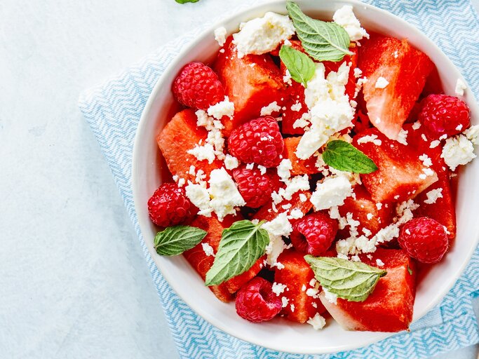 Wassermelonenslata mit Minze, Himbeeren und Feta in einer weißen Schale auf einem blauen Tuch. Auf einem hellen Tisch mit zwei Holzgabeln an der rechten Seite. | © Gettyimages/ nerudol