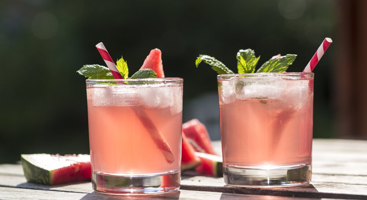 Zwei Gläser gefüllt mit rosafarbenem Wasser-Melonen-Hugo – Dekoriert mit Frucht, Minze und Strohalmen. 
