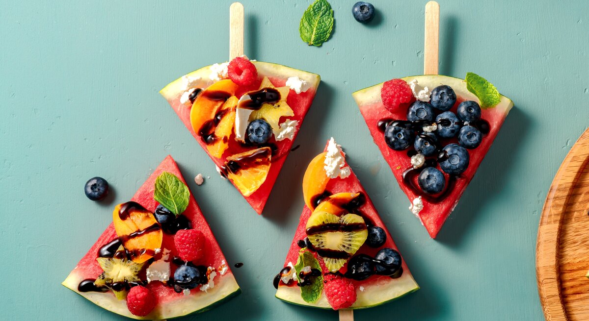Vier Stücken Wassermelone, belegt mit Obst auf einem grünem Tisch. Alles direkt von oben fotografiert.