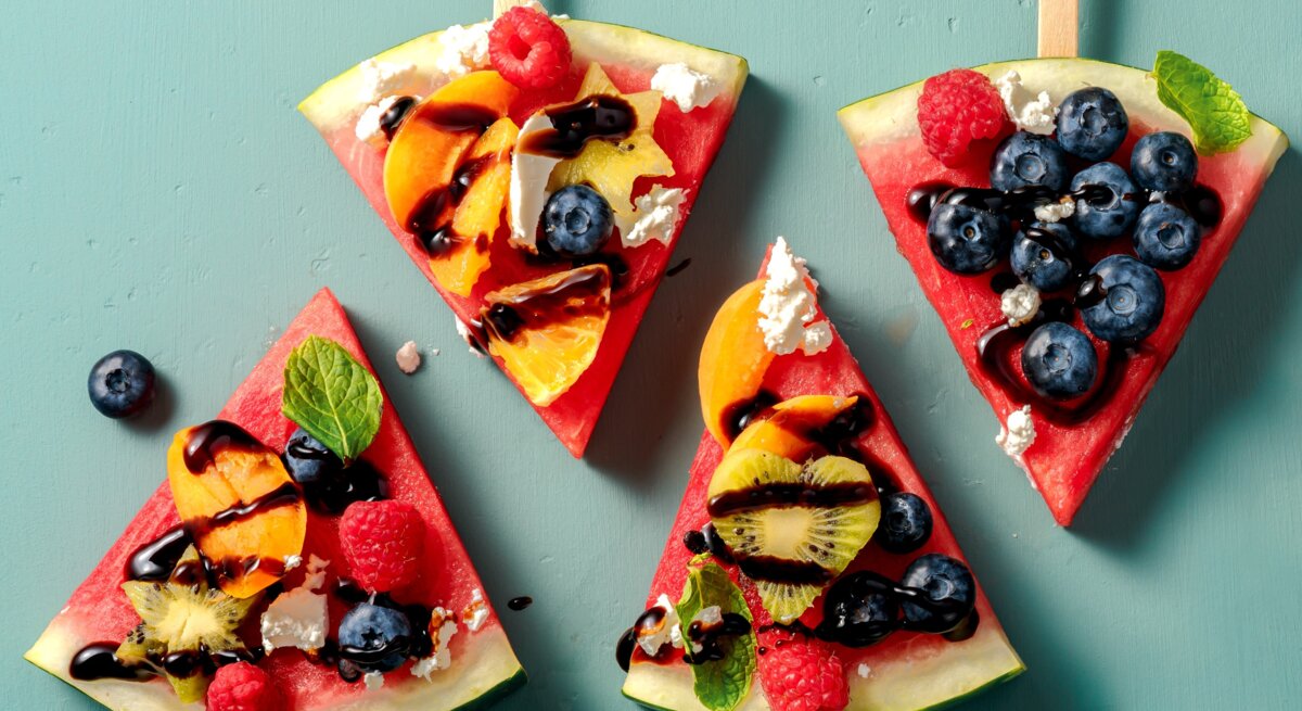Vier Stücken Wassermelone, belegt mit Obst auf einem grünem Tisch. Alles direkt von oben fotografiert.