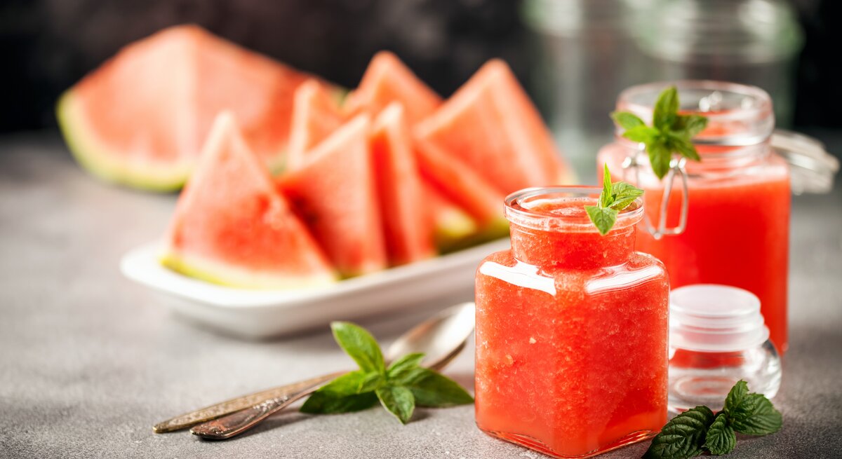 Zwei Gläser mit Wassermelonen-Minz-Gelee stehen auf einem grauen Untergrund. Daneben liegen Minz-Blätter und Löffel. Eine Schale mit Melonenstücken liegt im Hintergrund.