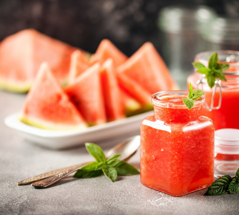 Zwei Gläser mit Wassermelonen-Minz-Gelee stehen auf einem grauen Untergrund. Daneben liegen Minz-Blätter und Löffel. Eine Schale mit Melonenstücken liegt im Hintergrund. | © Shutterstock / NatalyaBond