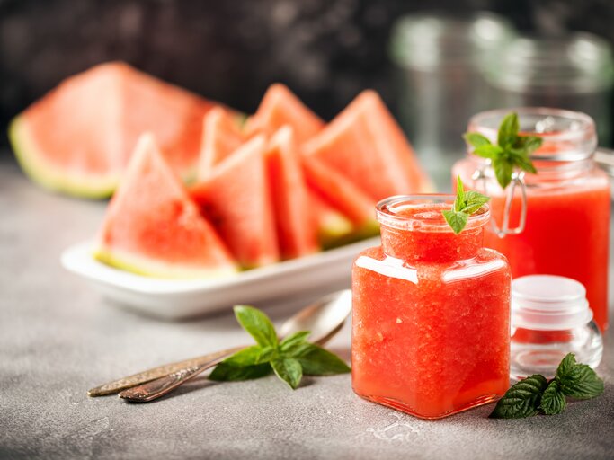 Zwei Gläser mit Wassermelonen-Minz-Gelee stehen auf einem grauen Untergrund. Daneben liegen Minz-Blätter und Löffel. Eine Schale mit Melonenstücken liegt im Hintergrund. | © Shutterstock / NatalyaBond