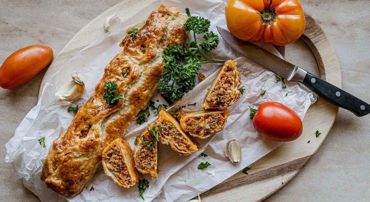 Hackfleisch-Blätterteig-Rolle auf einem Holzbrett. Daneben ein Messer und mehrere Tomaten. 