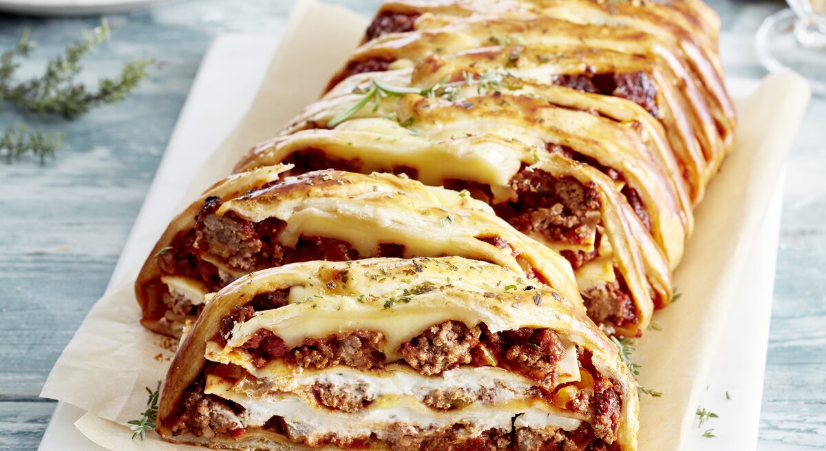 Ein Lasagne-Blätterzopf auf einem weißen Teller auf hellblauem Tisch.