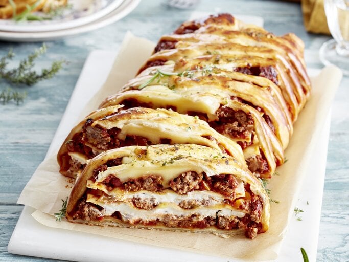Ein Lasagne-Blätterzopf auf einem weißen Teller auf hellblauem Tisch. | © EAT CLUB/Isabell Triemer
