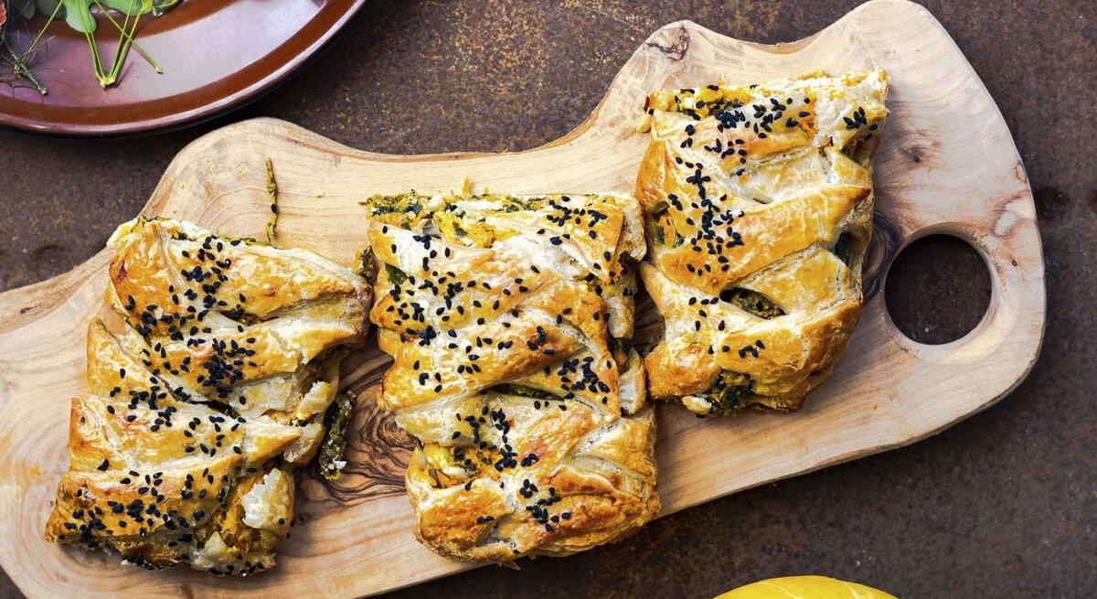 Auf einem Schneidebrett liegt Blätterteiggebäck gefüllt mit Kürbis, Spinat und Feta. Es ist mit Sesam bestreut.