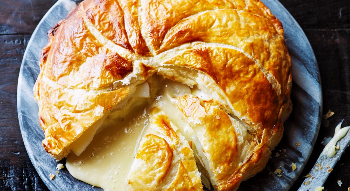 Ein großer Blätterteig-Pie mit Kartoffeln und Camembert auf einer blau-weißen, runden Scheibe, angeschnitten, sodass der geschmolzene Camembert hinausläuft, ein Stück fehlt, daneben ein Messer, von oben. 