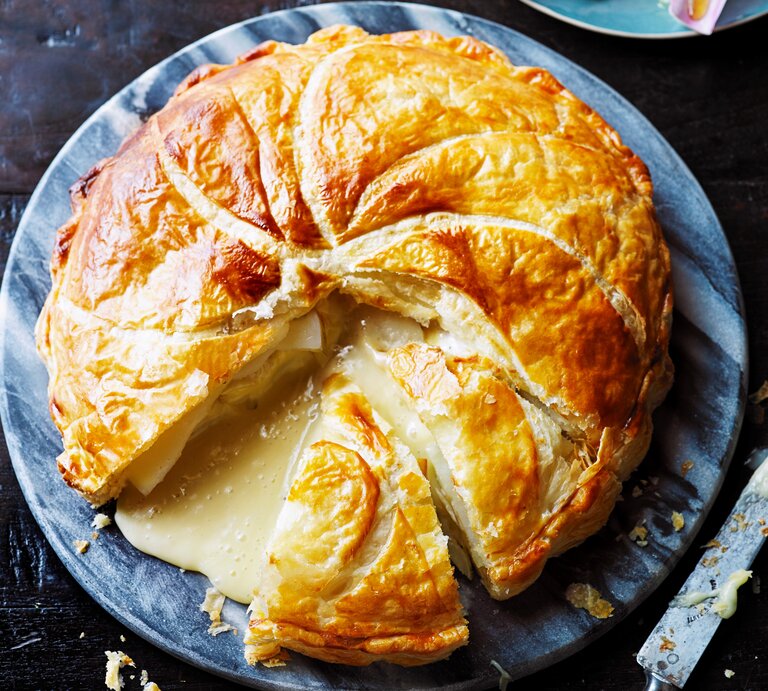 Ein großer Blätterteig-Pie mit Kartoffeln und Camembert auf einer blau-weißen, runden Scheibe, angeschnitten, sodass der geschmolzene Camembert hinausläuft, ein Stück fehlt, daneben ein Messer, von oben.  | © StockFood / Immediate Media / Good Food