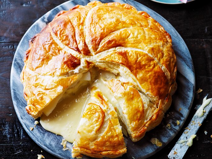 Ein großer Blätterteig-Pie mit Kartoffeln und Camembert auf einer blau-weißen, runden Scheibe, angeschnitten, sodass der geschmolzene Camembert hinausläuft, ein Stück fehlt, daneben ein Messer, von oben.  | © StockFood / Immediate Media / Good Food