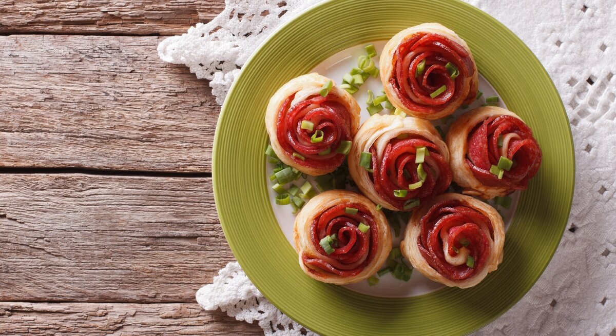 Mehre Salami-Blätterteig-Rosen auf einem weißen Teller mit grünem Rand und rustikalem Holztisch. Darunter noch eine weiße Tischdecke und alles direkt von oben fotografiert.