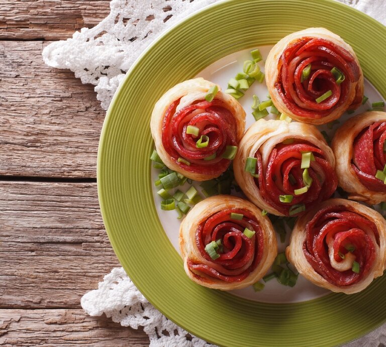 Mehre Salami-Blätterteig-Rosen auf einem weißen Teller mit grünem Rand und rustikalem Holztisch. Darunter noch eine weiße Tischdecke und alles direkt von oben fotografiert. | © Shutterstock/ Sergii Koval