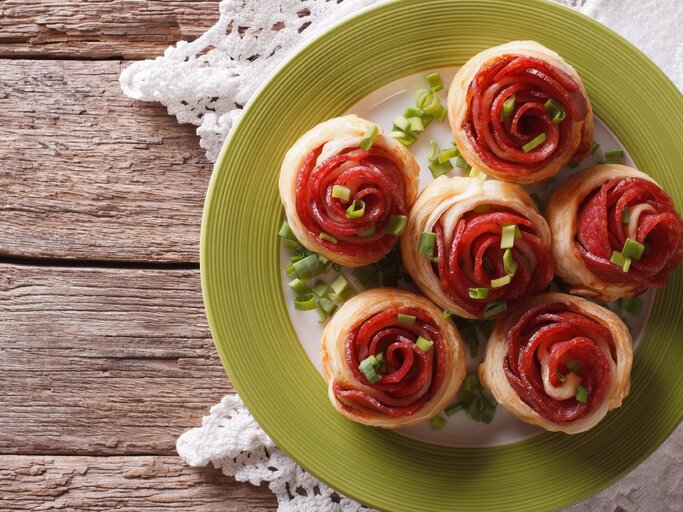 Mehre Salami-Blätterteig-Rosen auf einem weißen Teller mit grünem Rand und rustikalem Holztisch. Darunter noch eine weiße Tischdecke und alles direkt von oben fotografiert. | © Shutterstock/ Sergii Koval