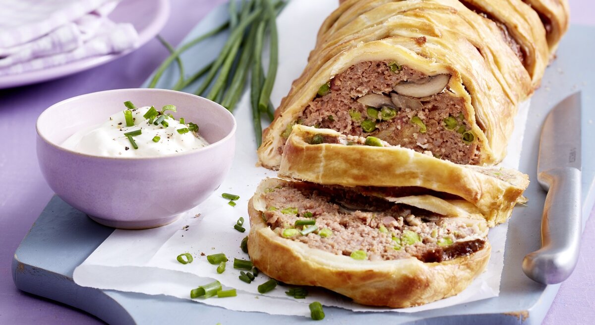 Hackbraten im Blätterteigmantel mit einer Füllung aus Hackfleisch, Pilzen, Erbsen und Schinken. Angeschnitten serviert auf einem Holzbrett und serviert mit frischem Schnittlauchschmand. 