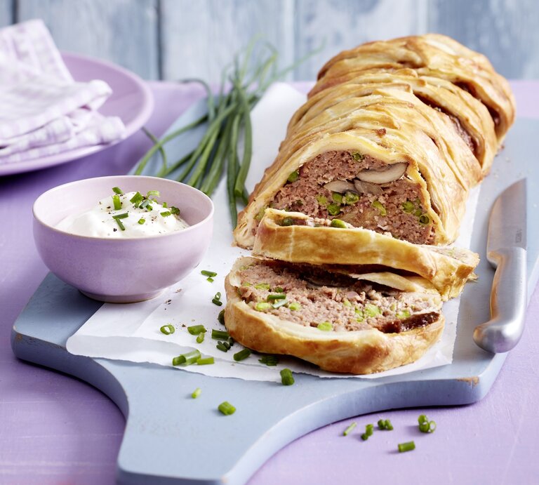 Hackbraten im Blätterteigmantel mit einer Füllung aus Hackfleisch, Pilzen, Erbsen und Schinken. Angeschnitten serviert auf einem Holzbrett und serviert mit frischem Schnittlauchschmand.  | © EAT CLUB / Isabell Triemer