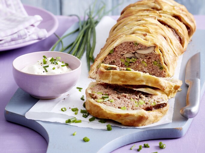 Hackbraten im Blätterteigmantel mit einer Füllung aus Hackfleisch, Pilzen, Erbsen und Schinken. Angeschnitten serviert auf einem Holzbrett und serviert mit frischem Schnittlauchschmand.  | © EAT CLUB / Isabell Triemer