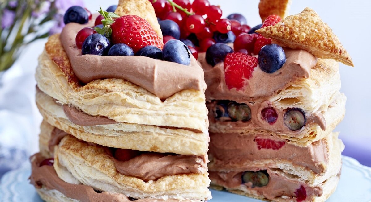 Auf einem Teller steht eine Schoko-Beeren-Torte. Sie hat mehrere Schichten Creme und ist mit verschiedenen Beeren dekoriert.