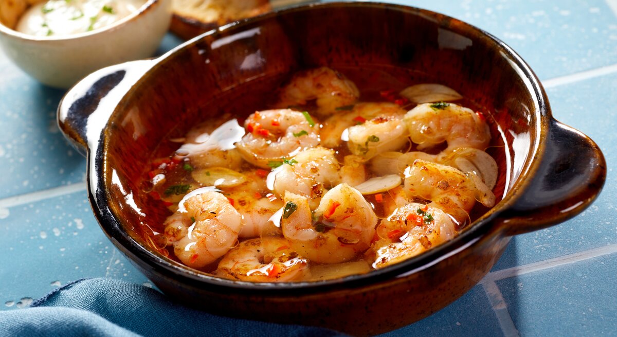 Draufsicht, schräg: Eine Tonschüssel mit Shrimps al ajillo steht auf blauen Kacheln mit Fugen. Daneben liegen Weißbrotscheiben und einen Schüssel mit einer hellen Creme.