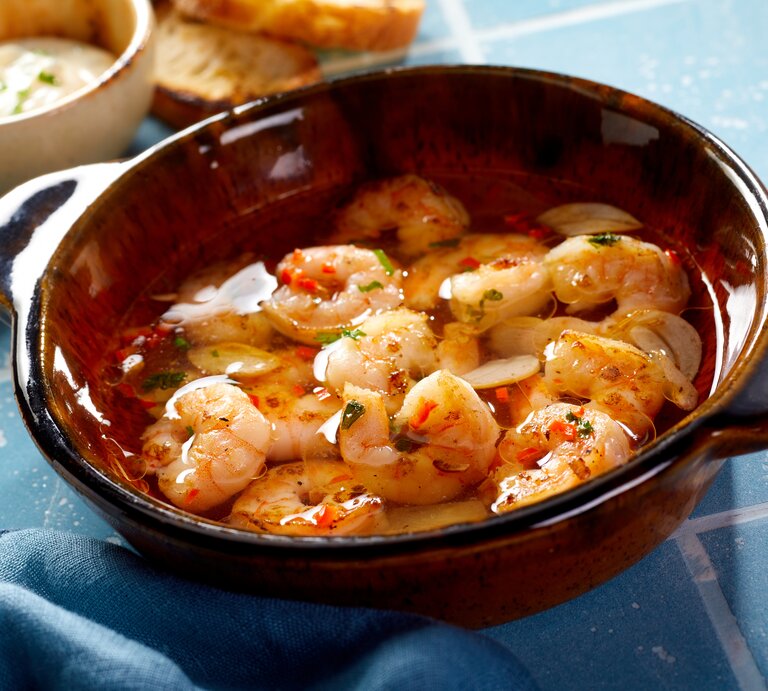 Draufsicht, schräg: Eine Tonschüssel mit Shrimps al ajillo steht auf blauen Kacheln mit Fugen. Daneben liegen Weißbrotscheiben und einen Schüssel mit einer hellen Creme. | © Shutterstock / stockcreations