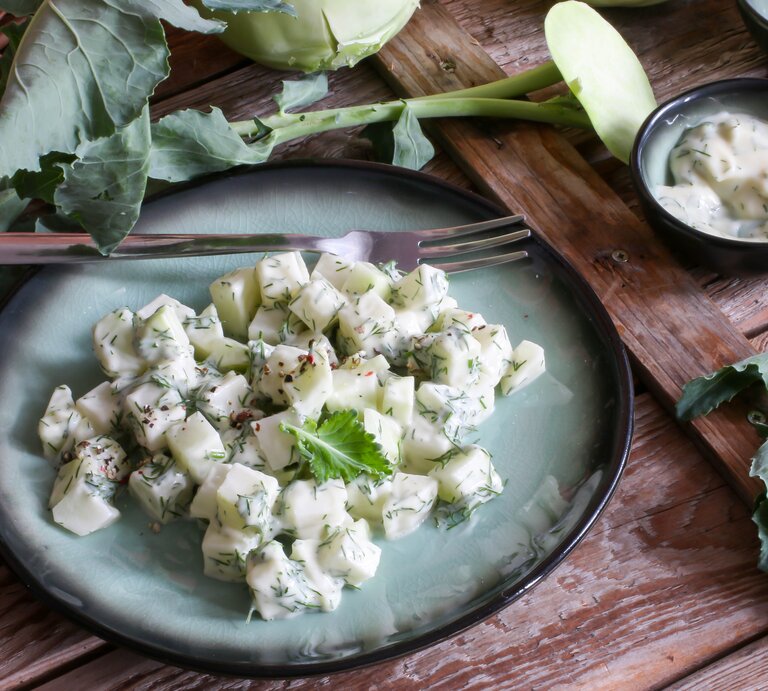 Schräge Draufsicht: ein blau-schwarzer Teller Kohlrabi-Joghurt-Salat steht auf einem dunklen Holztisch. Drum herum liegt frischer Kohlrabi. Und danaben ist ein Schälchen mit einer cremigen Soße. | © Shutterstock / Foxxy63