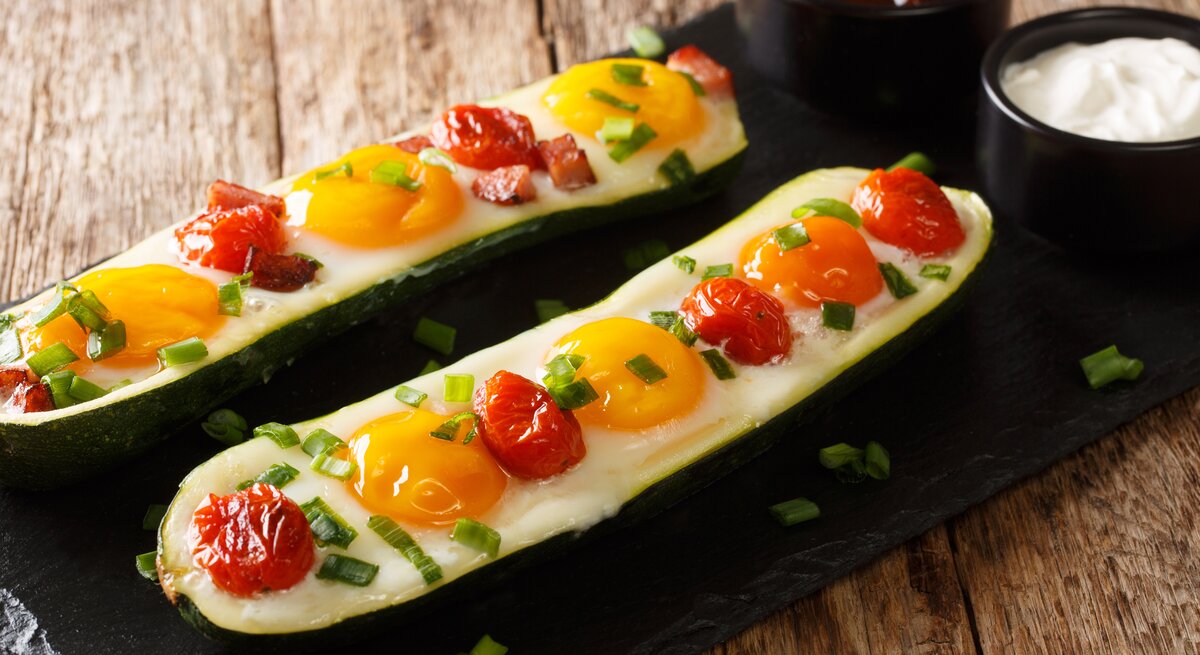 Zwei Spiegelei-Zucchini-Boote mit Kirschtomaten, eins mit Speck und beide mit Frühlingszwiebeln garniert auf einem schwarzen Brett neben zwei Schüsseln mit Soßen, vor einem Holzhintergrund. 