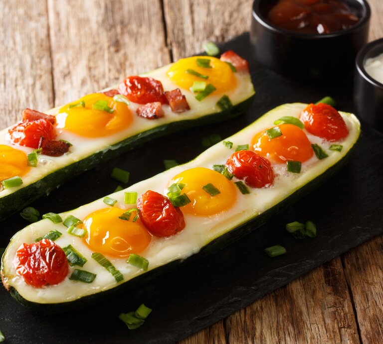 Zwei Spiegelei-Zucchini-Boote mit Kirschtomaten, eins mit Speck und beide mit Frühlingszwiebeln garniert auf einem schwarzen Brett neben zwei Schüsseln mit Soßen, vor einem Holzhintergrund.  | © Getty Images/ALLEKO