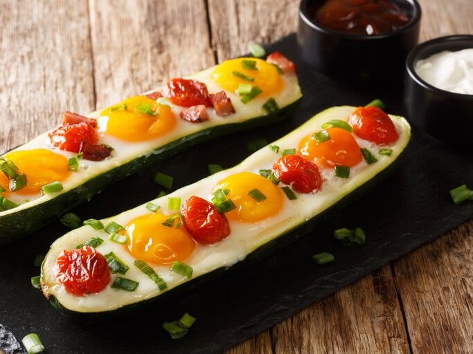 Zwei Spiegelei-Zucchini-Boote mit Kirschtomaten, eins mit Speck und beide mit Frühlingszwiebeln garniert auf einem schwarzen Brett neben zwei Schüsseln mit Soßen, vor einem Holzhintergrund.  | © Getty Images/ALLEKO