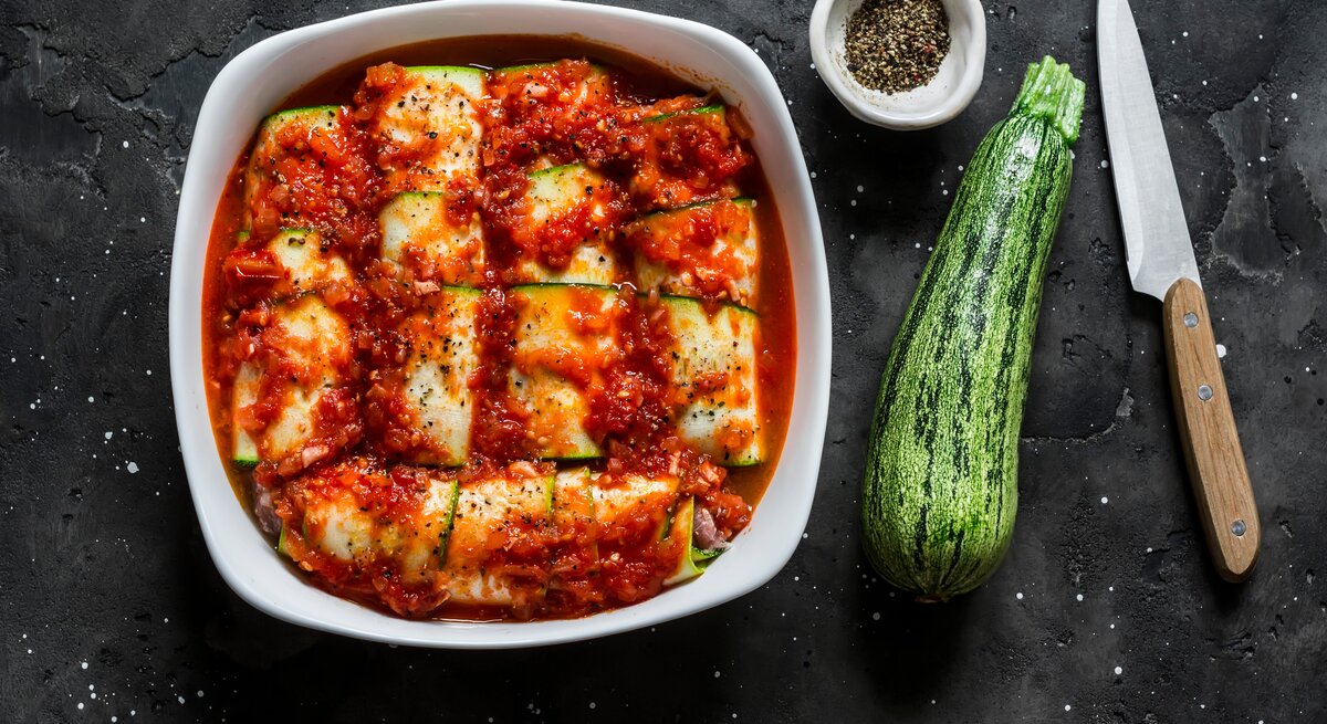 Low Carb Zucchini-Cannelloni mit Spinat-Ricotta-Füllung und fruchtiger Tomatensoße in Auflaufform auf schwarzem Untergrund. Rechts kleine Schale mit buntem Pfeffer. Daneben eine Zucchini und ein Messer mit Holzgriff. Aufnahme aus der Vogelperspektive. 