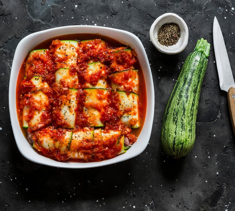 Low Carb Zucchini-Cannelloni mit Spinat-Ricotta-Füllung und fruchtiger Tomatensoße in Auflaufform auf schwarzem Untergrund. Rechts kleine Schale mit buntem Pfeffer. Daneben eine Zucchini und ein Messer mit Holzgriff. Aufnahme aus der Vogelperspektive.  | © shutterstock / Kiian Oksana