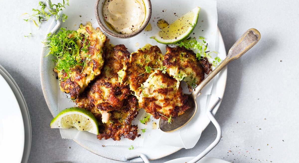 Mehrere Hüttenkäse-Taler mit Zucchini auf einem weißen Teller mit Limette, Kresse und einem hellen Dip auf. Alles auf einem hellen Tisch, direkt von oben fotografiert.