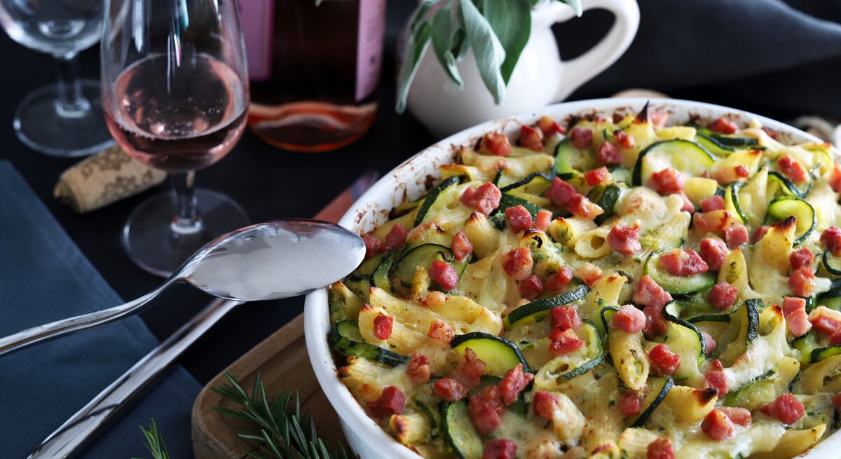 Würziger Zucchini-Nudelauflauf mit saftigen Speckwürfeln bestreut und überbacken mit Käse, serviert in einer runden, weißen Auflaufform. Daneben stehen Gläser und ein wenig Dekoration. 