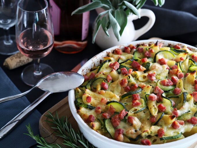 Würziger Zucchini-Nudelauflauf mit saftigen Speckwürfeln bestreut und überbacken mit Käse, serviert in einer runden, weißen Auflaufform. Daneben stehen Gläser und ein wenig Dekoration.  | © Shutterstock/Grazziela
