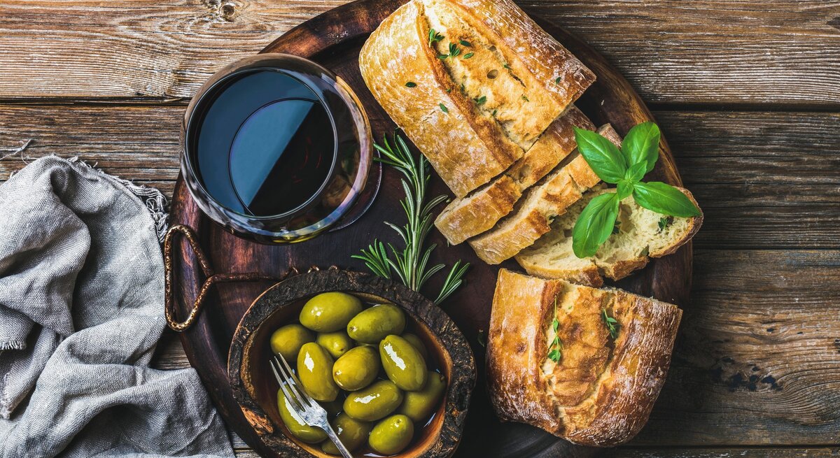 Schnelles Olivenbrot auf dunklem Holzteller mit Glas Rotwein und Kräutern und Oliven auf Holztisch. Daneben graue Stoffserviette. Aufnahme aus der Vogelperspektive.
