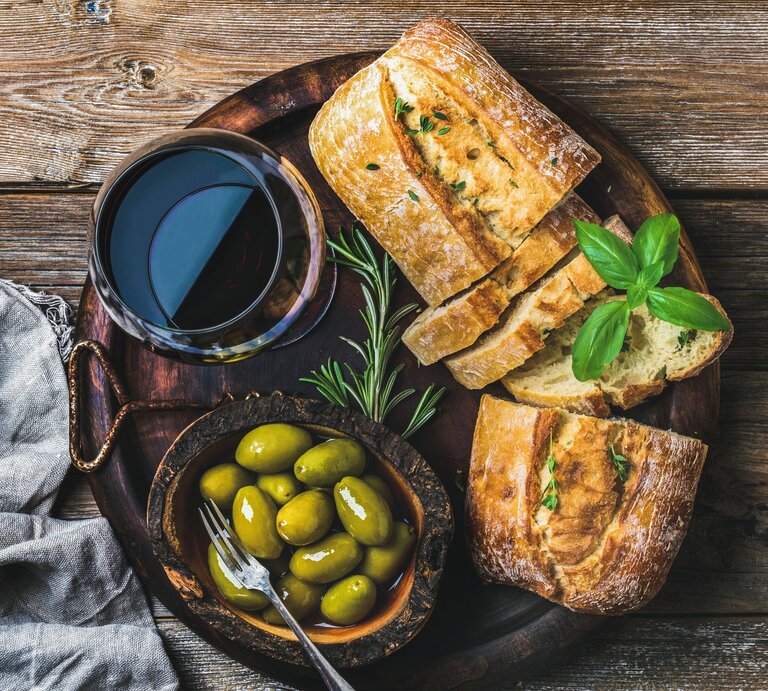 Schnelles Olivenbrot auf dunklem Holzteller mit Glas Rotwein und Kräutern und Oliven auf Holztisch. Daneben graue Stoffserviette. Aufnahme aus der Vogelperspektive. | © Getty Images / Foxys_forest_manufacture