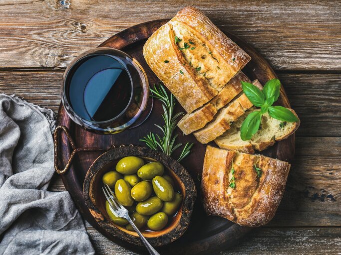 Schnelles Olivenbrot auf dunklem Holzteller mit Glas Rotwein und Kräutern und Oliven auf Holztisch. Daneben graue Stoffserviette. Aufnahme aus der Vogelperspektive. | © Getty Images / Foxys_forest_manufacture