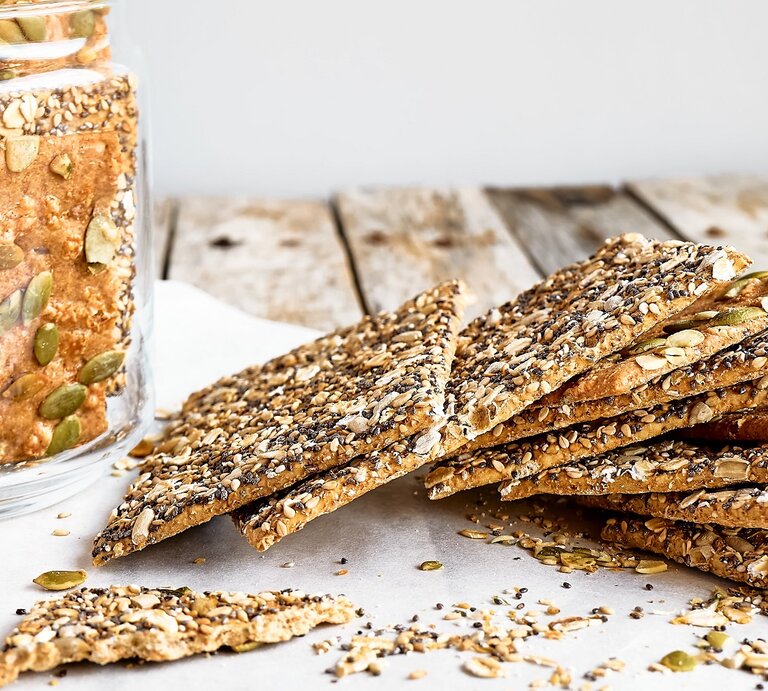 Mehrere Knäckebrote auf einem Holztisch mit Backpapier und einem Glas mit dem Knäckebrot. | © Shutterstock/ Caterina Trimarchi