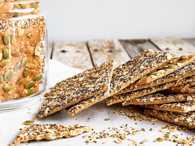 Mehrere Knäckebrote auf einem Holztisch mit Backpapier und einem Glas mit dem Knäckebrot. | © Shutterstock/ Caterina Trimarchi