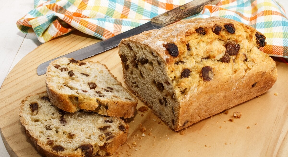 Frontalsicht: Auf einem Holzbrettchen liegt ein angeschnittenes Rosinenbrot und daneben ein Messer mit einem Holzschaft. Daneben liebt ein Küchenhandtuch.