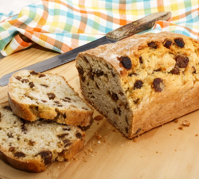 Frontalsicht: Auf einem Holzbrettchen liegt ein angeschnittenes Rosinenbrot und daneben ein Messer mit einem Holzschaft. Daneben liebt ein Küchenhandtuch. | © Shutterstock / Joerg Beuge
