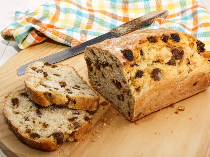 Frontalsicht: Auf einem Holzbrettchen liegt ein angeschnittenes Rosinenbrot und daneben ein Messer mit einem Holzschaft. Daneben liebt ein Küchenhandtuch. | © Shutterstock / Joerg Beuge