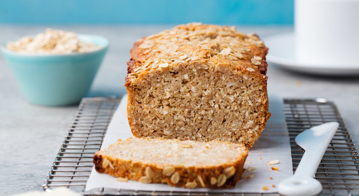 Bananenbrot mit Haferflocken auf Kuchenrost, dahinter eine blaue Schüssel mit Haferflocken.