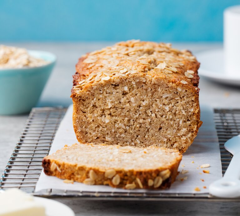 Bananenbrot mit Haferflocken auf Kuchenrost, dahinter eine blaue Schüssel mit Haferflocken. | © Getty Images/ AnnaPustynnikova