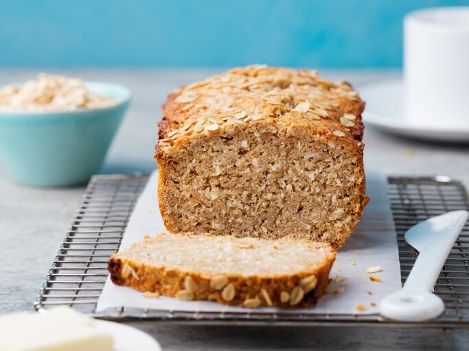Bananenbrot mit Haferflocken auf Kuchenrost, dahinter eine blaue Schüssel mit Haferflocken. | © Getty Images/ AnnaPustynnikova