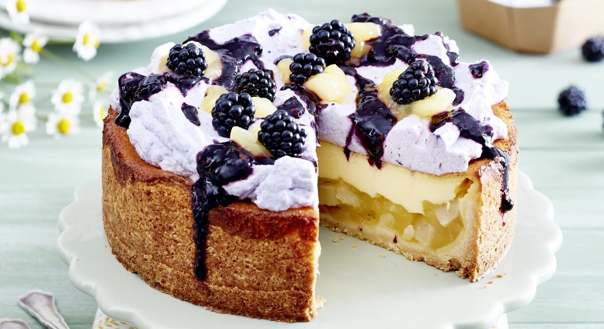 Auf einem Teller steht eine Birnen-Brombeer-Schmandtorte. Sie ist mit einer lila Creme verziert und mit Früchten bedeckt. Darüber wurde eine Brombeersoße gegeben. 