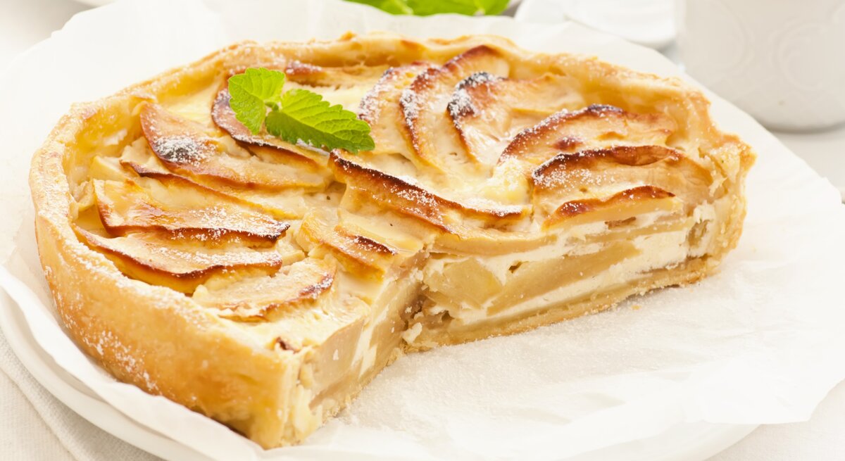 Die angeschnittene Birnen-Schmand-Tarte auf einem weißen Teller mit Backpapier. Alles auf hellem Unter- und Hintergrund fotografiert mit einem weißen Kaffeegedeck.