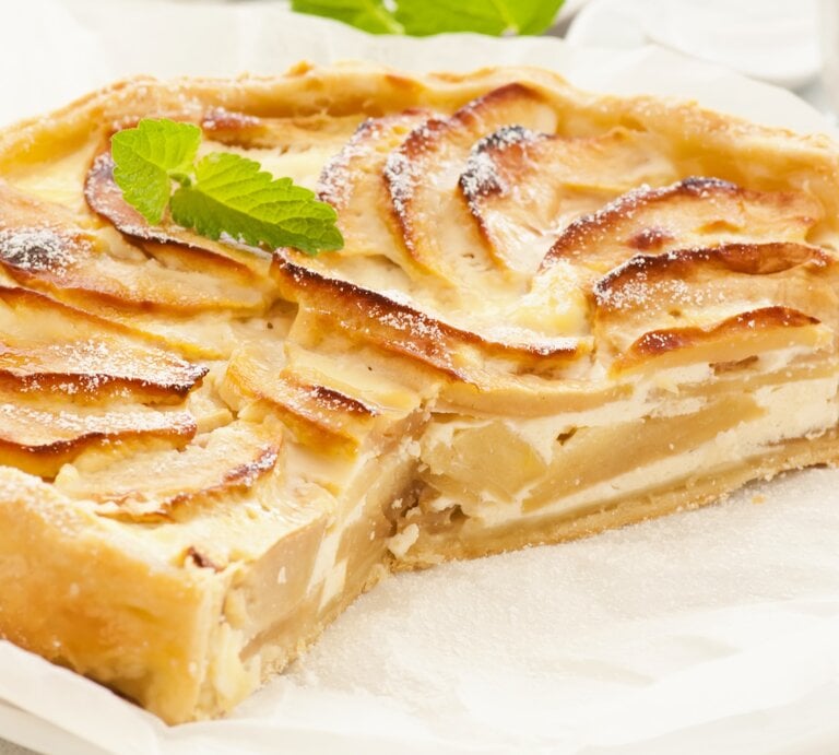 Die angeschnittene Birnen-Schmand-Tarte auf einem weißen Teller mit Backpapier. Alles auf hellem Unter- und Hintergrund fotografiert mit einem weißen Kaffeegedeck. | © Shutterstock/ hlphoto