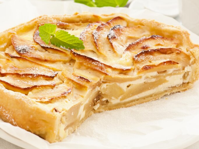 Die angeschnittene Birnen-Schmand-Tarte auf einem weißen Teller mit Backpapier. Alles auf hellem Unter- und Hintergrund fotografiert mit einem weißen Kaffeegedeck. | © Shutterstock/ hlphoto