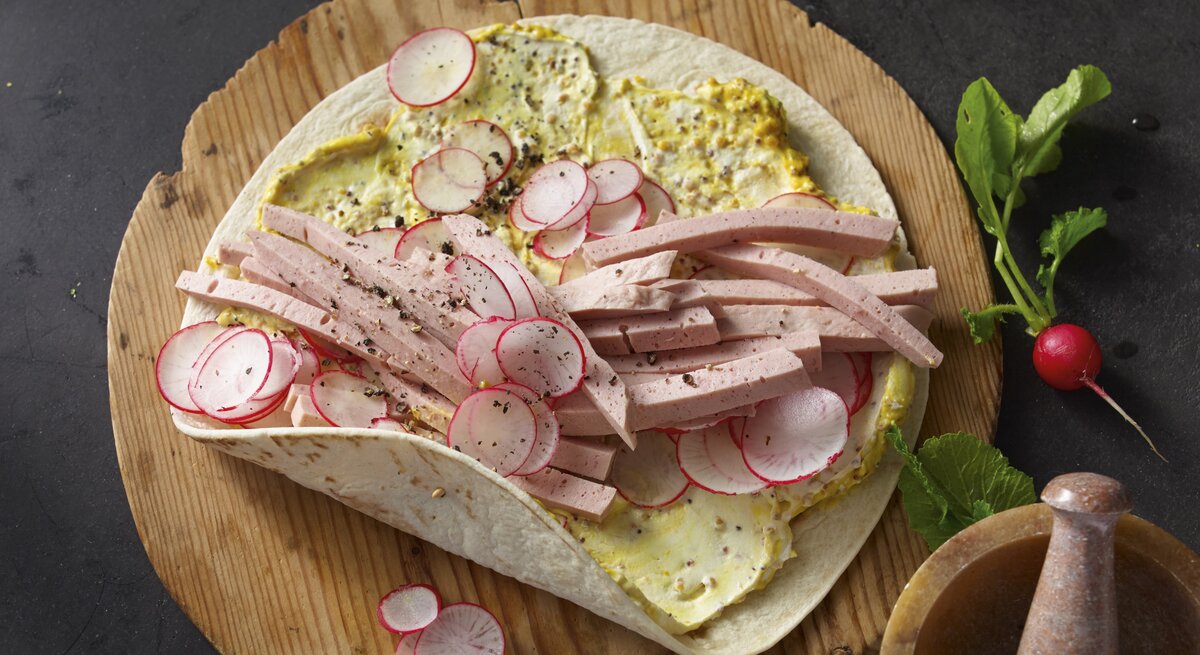 Bayern-Wraps mit Leberkäse, Senfcreme und Radieschen, garniert mit frischem Pfeffer und serviert auf einem runden Holzbrett. 